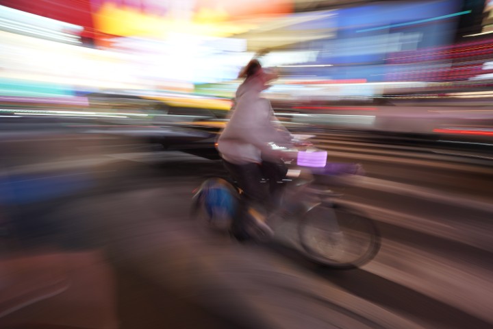 A night photo of a person riding a bike. The picture shows a lot of motion blur as the photographer was following the bike passing by. The background is blurry and colorful, and the bike is partly in focus, giving a sense of speed.