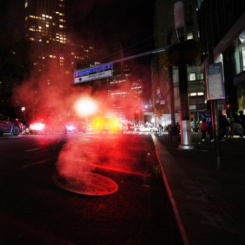 Street of NYC, at night. A manhole, with smoke coming out, is visible in the foreground.